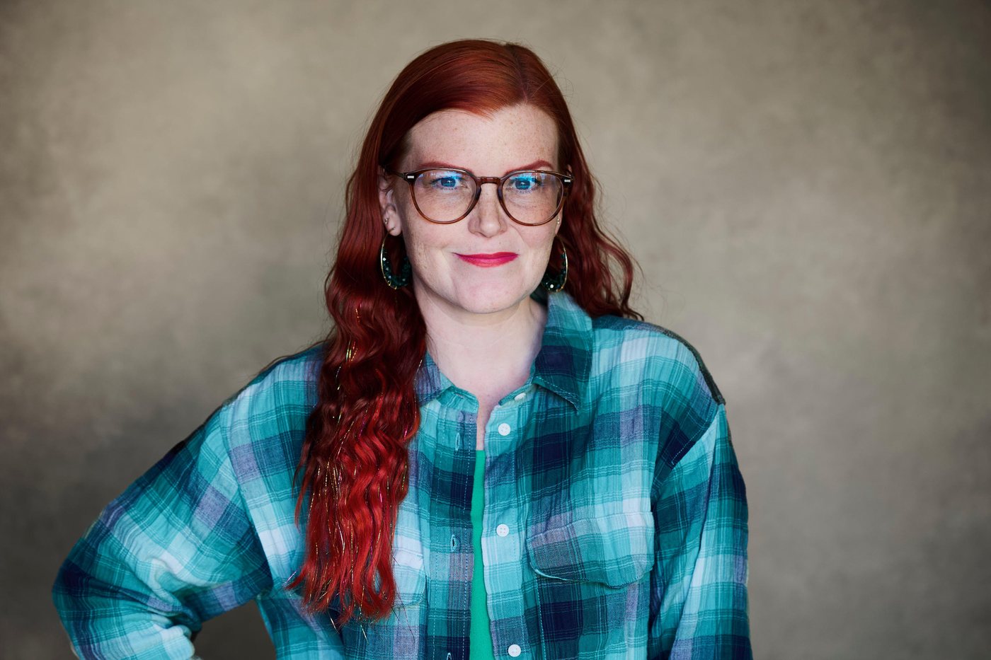 Jackie Nash in a teal plaid shirt and tortoiseshell glasses, photographed against a grey studio backdrop.